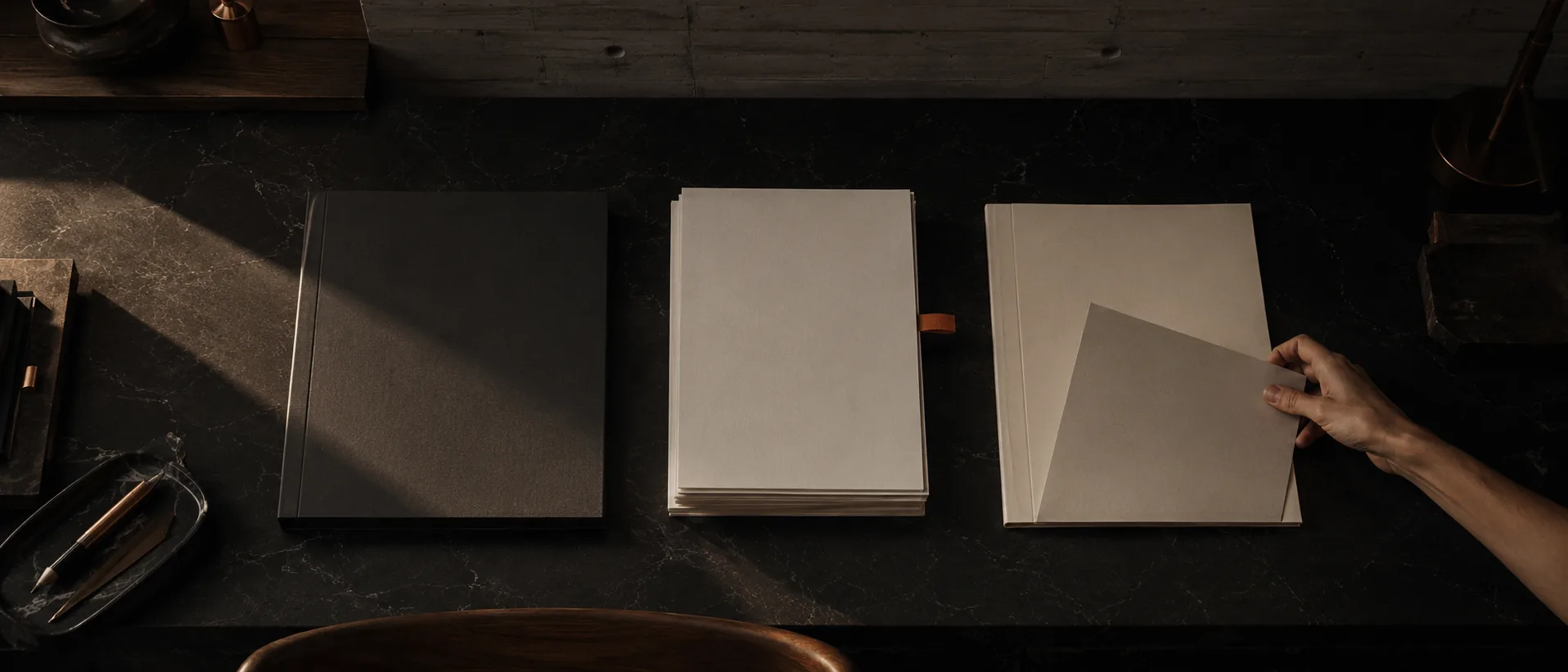 Overhead view of a dark stone desk with three document sets in a row: a black portfolio, a stack of white pages, and a folder a hand is opening